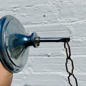 May include: A chrome-plated ceiling canopy with a rod and a rusty chain. The canopy has a circular base and a decorative, rounded center. The chain is attached to the rod and hangs down. The background is a white brick wall.