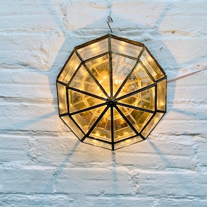 Könnte beinhalten: Eine achteckige Wandleuchte im Vintage-Stil mit geometrischem Design. Die Leuchte hat einen messingfarbenen Rahmen und klare Glasscheiben, die von warm getönten Lichtern beleuchtet werden. Sie ist an einer weißen Backsteinmauer befestigt.