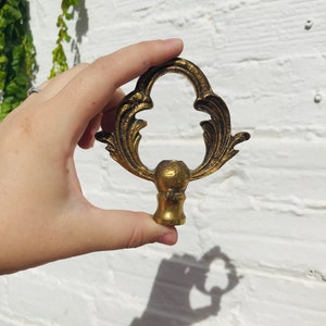 Puede incluir: Un remate de metal dorado con un diseño decorativo y ornamentado. El remate tiene una base redonda y una parte superior en forma de corazón.