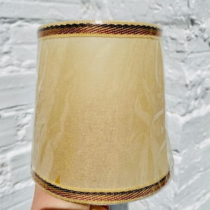 May include: A beige lampshade with a brown, gold, and black trim. The lampshade is made of fabric and has a slightly textured surface.