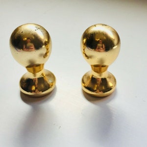 Op de afbeelding: Twee goudkleurige, bolvormige kastknoppen. Elke knop heeft een ronde basis en een korte, gebogen steel die verbinding maakt met de bolvormige bovenkant. De knoppen zijn reflecterend, met een glanzende, metalen afwerking.