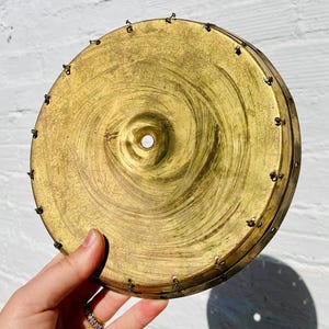 May include: A vintage brass-colored metal object with a circular shape. The object has a central hole and is adorned with small rivets around the edge. The surface shows a spiral pattern and signs of wear, suggesting age. The item is held by a hand.