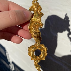 Könnte beinhalten: Ein verzierter, goldfarbener Metall-Dekorzug mit einem runden Loch und einem geschwungenen Design.