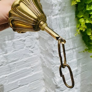 Op de afbeelding: Een vintage messing plafondarmatuurkap met een decoratief, gegroefd ontwerp. De kap is bevestigd aan een messing staaf en ketting. De armatuur heeft een warme goudkleur en staat tegen een witte bakstenen muur.