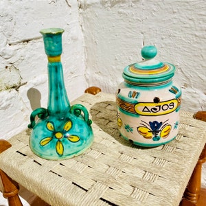 May include: Two ceramic containers with floral designs. The larger container is white with blue and yellow accents and says "AJOS" on the front. The smaller container is teal with yellow flowers and is a candle holder.