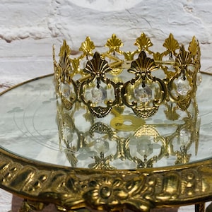 Puede incluir: Una corona de metal dorado con detalles de cristal transparente. La corona está sobre una superficie de vidrio con una base de metal dorado.