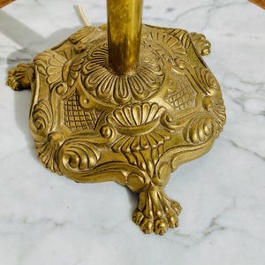 Könnte beinhalten: Eine goldene Metall-Lampenbasis mit einem verzierten Design. Die Basis hat eine runde Oberseite und drei Beine mit komplizierten Details.