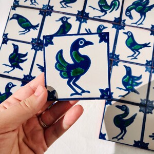 May include: A close-up of a blue and green bird tile. The tile is white with a blue border and a green and blue bird design. The bird has a long neck and a long tail. The tile is part of a set of tiles with different bird designs.