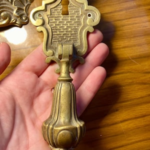 Puede incluir: Tirador de armario antiguo de latón con detalles ornamentados. El tirador presenta un diseño de ojo de cerradura y un asa decorativa en forma de campana. El latón tiene una pátina cálida y envejecida, lo que sugiere artesanía vintage.