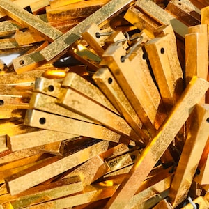 Op de afbeelding: Een close-up van een stapel rechthoekige, goudkleurige metalen stukken. Elk stuk heeft een klein gat dat door één uiteinde is geboord en een draadlus. De stukken zijn gestapeld en overlappen elkaar, waardoor een textuur ontstaat.