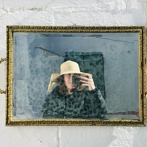 May include: A rectangular antique mirror with a gold-colored ornate frame and handles. The mirror reflects a person wearing a hat and jacket. The mirror is hanging on a white brick wall.