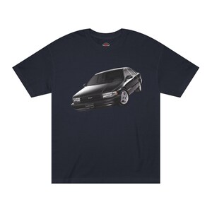 Könnte beinhalten: Marineblaues T-Shirt mit einer Grafik eines schwarzen Autos. Das Auto hat ein schlankes Design mit silbernen Felgen und ist vor einem dunklen Hintergrund platziert. Das T-Shirt besteht aus einem weichen, bequemen Material.