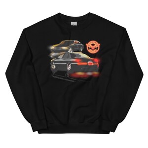 Könnte beinhalten: Schwarzes Sweatshirt mit einer Grafik von zwei schwarzen Autos mit leuchtenden Scheinwerfern und Rücklichtern. Ein neonorangefarbenes Emblem befindet sich oben rechts. Das Sweatshirt ist aus weichem Material.