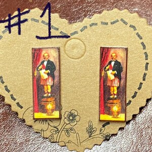 May include: A pair of rectangular stud earrings featuring a vintage illustration of a man standing on a barrel of dynamite. The man is dressed in a suit and holding a scroll, with a red curtain backdrop.