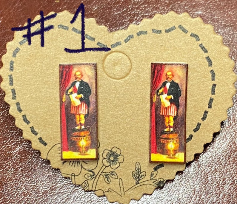 May include: A pair of rectangular stud earrings featuring a vintage illustration of a man standing on a barrel of dynamite. The man is dressed in a suit and holding a scroll, with a red curtain backdrop.