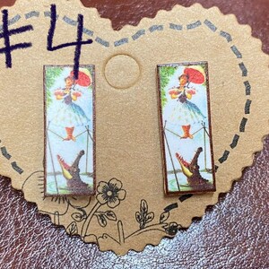 May include: A pair of rectangular earrings featuring a vintage illustration of a woman with a red umbrella and a crocodile. The earrings are set on a heart-shaped cardboard backing with the number "4" written in dark blue marker.