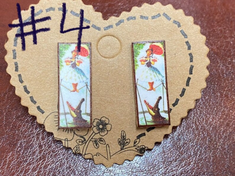 May include: A pair of rectangular earrings featuring a vintage illustration of a woman with a red umbrella and a crocodile. The earrings are set on a heart-shaped cardboard backing with the number "4" written in dark blue marker.