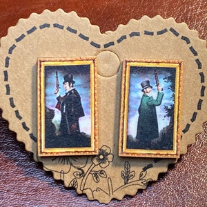 May include: A pair of stud earrings featuring miniature framed portraits of men holding rifles. The portraits have a vintage aesthetic with a gold frame and are set on a heart-shaped cardboard backing.