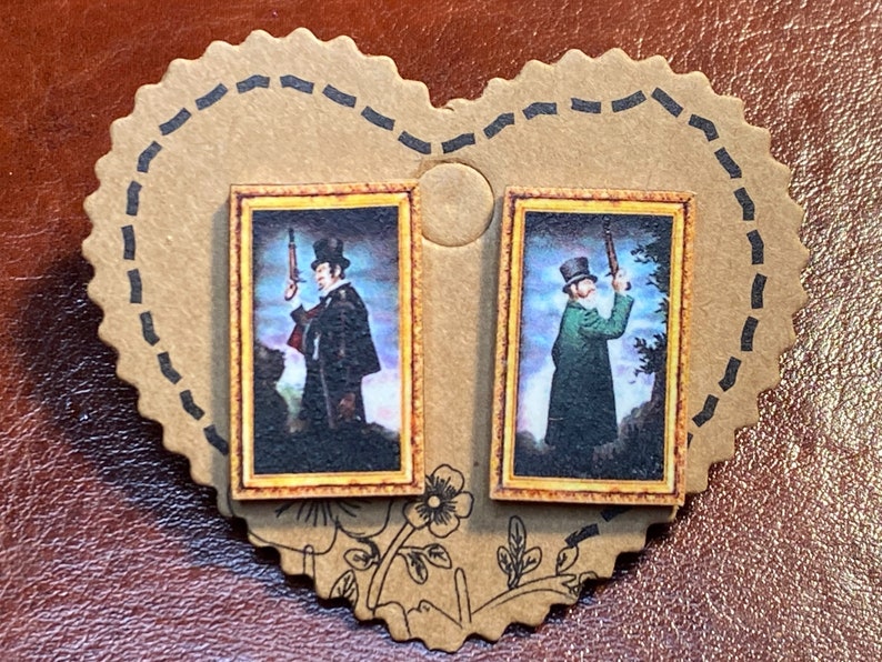 May include: A pair of stud earrings featuring miniature framed portraits of men holding rifles. The portraits have a vintage aesthetic with a gold frame and are set on a heart-shaped cardboard backing.