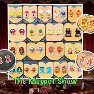 May include: A display of colorful Muppet Show character earrings. The earrings are arranged on display cards, featuring various characters like Kermit, Miss Piggy, and Fozzie Bear. The text "The Muppet Show" is visible at the bottom.