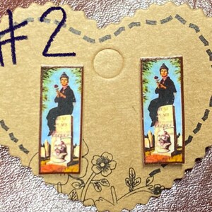 May include: A pair of rectangular earrings featuring a vintage-style image of a person holding a rose, seated on a tombstone. The image is set against a blue sky background. The earrings are displayed on a heart-shaped, tan card.