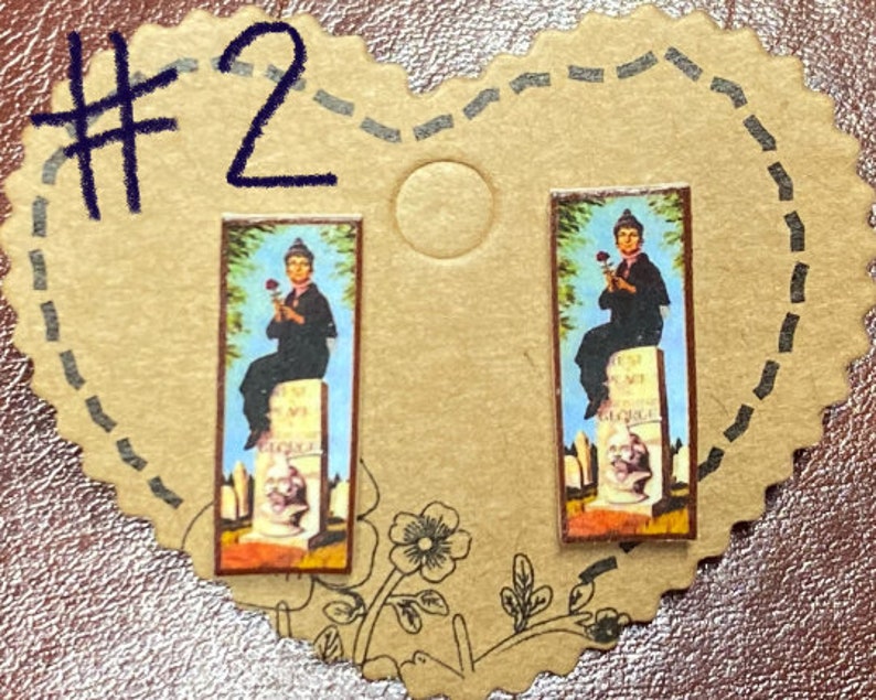 May include: A pair of rectangular earrings featuring a vintage-style image of a person holding a rose, seated on a tombstone. The image is set against a blue sky background. The earrings are displayed on a heart-shaped, tan card.