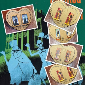May include: A collection of handmade earrings featuring miniature portraits of figures from a haunted mansion. The earrings are mounted on heart-shaped cardboard with decorative edges. The background features a green illustration of a haunted house and ghostly figures.