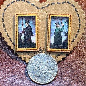 May include: A pair of small, rectangular earrings featuring miniature framed portraits of men holding rifles. The portraits have a vintage aesthetic with gold-colored frames. The earrings are displayed on a brown, heart-shaped card.