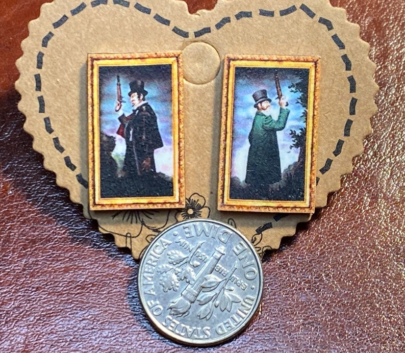 May include: A pair of small, rectangular earrings featuring miniature framed portraits of men holding rifles. The portraits have a vintage aesthetic with gold-colored frames. The earrings are displayed on a brown, heart-shaped card.