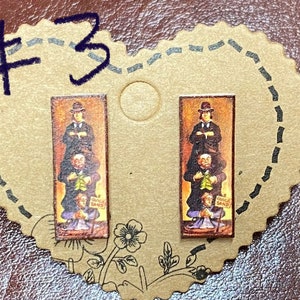 May include: A pair of rectangular stud earrings featuring a vintage-style image of a man standing on top of three other men. The image includes the text "QUICKSAND". The earrings are set on a heart-shaped card with the number "3" written on it.