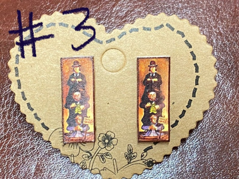 May include: A pair of rectangular stud earrings featuring a vintage-style image of a man standing on top of three other men. The image includes the text "QUICKSAND". The earrings are set on a heart-shaped card with the number "3" written on it.