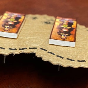 May include: A pair of rectangular stud earrings. Each earring features a colorful, detailed image with a white border. The earrings are set on a tan, scalloped surface with a dotted black line around the edge.