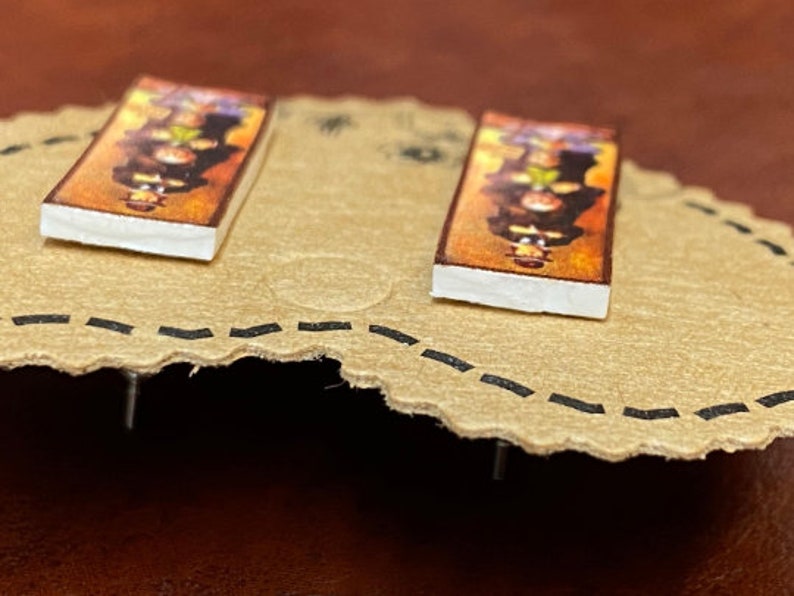 May include: A pair of rectangular stud earrings. Each earring features a colorful, detailed image with a white border. The earrings are set on a tan, scalloped surface with a dotted black line around the edge.