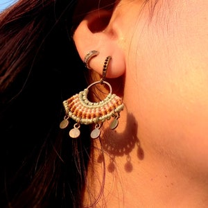 May include: A pair of earrings with a woven, fan-shaped design in shades of pink, white, and brown. The earrings have small, round, metallic charms hanging from the bottom.