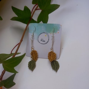 May include: A pair of dangle earrings with two leaf charms. The top leaf is a mustard yellow colour and the bottom leaf is a dark green colour. The earrings are on a white card with the word "Rbe" written on it.