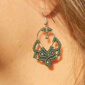 May include: Green and silver dangle earrings with a delicate, intricate design. The earrings are made of a knotted cord and have a small silver drop pendant.