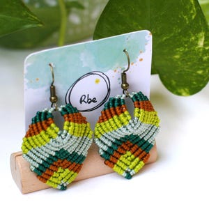 Op de afbeelding: Handgemaakte macramé oorbellen in een bladmotief. De oorbellen hebben een combinatie van groene, gele, oranje en witte draden. De oorbellen worden gepresenteerd op een houten standaard met de tekst "Rbe" in een cirkel.