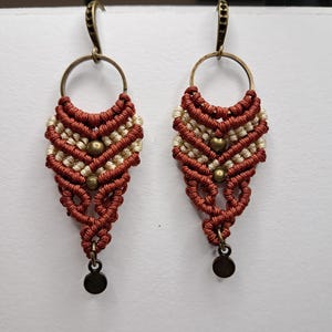 May include: A pair of brown and white macrame earrings with gold beads and a bronze hook. The earrings are hanging from a white background.