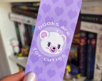 Puede incluir: Un marcapáginas rectangular de color morado con un patrón repetitivo de corazones pixelados. En el centro, un diseño circular contiene una cara de oso blanco pixelada con orejas rosas y el texto "books are for cuties" arqueado alrededor.