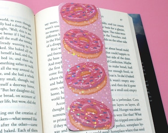 Puede incluir: Un marcapáginas de donut pixelado rosa y blanco con chispas de colores. El marcapáginas tiene un acabado brillante.