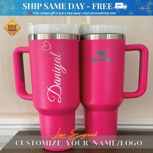 May include: Two pink stainless steel travel mugs with lids and handles. One mug is engraved with the name "Daniyal" and the other mug is engraved with the Stanley logo. The text "Laser Engraved" is below the mugs. The text "Customize Your Name/Logo" is below the engraved text.