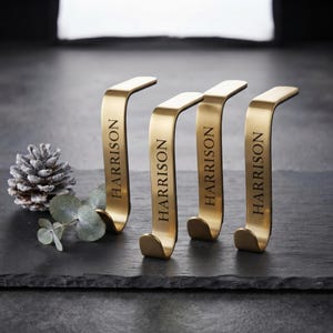 May include: Four gold-toned metal stocking holders with a curved, hook-shaped design. Each holder features the name HARRISON engraved in black capital letters vertically along the front surface of the metal stand.