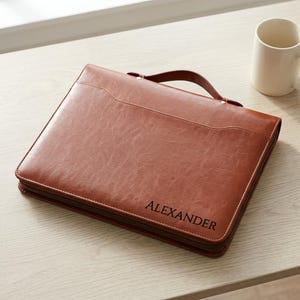 May include: A brown leather-like zippered portfolio case with a top handle and a curved front pocket. The name ALEXANDER is embossed in black capital letters on the lower right corner of the front cover, which features a textured finish.