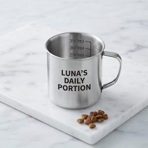 May include: A silver stainless steel measuring cup with a handle, featuring the black text LUNA'S DAILY PORTION and internal measurement markings up to 350ml. A small pile of brown dry pet food kibble sits next to the cup on a white surface.