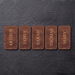 May include: Five rectangular brown leather labels with rounded corners, each featuring a name engraved in a light tan font. The names listed from left to right are ARTHUR, JAMES, SARAH, THOMAS, and MARY, all displayed in a vertical orientation.