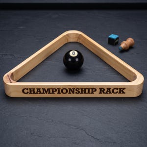 May include: A light-colored wooden triangular billiard ball rack with the text "CHAMPIONSHIP RACK" engraved on the front side. Inside the rack sits a black billiard ball with the number 8. A blue chalk cube and a wooden cue tip tool are nearby.