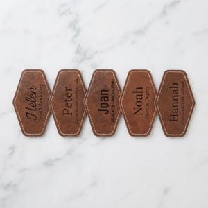 May include: Five brown leather-like motel-style key tags with white stitched edges. Each tag features a name and a font style: Helen with Great Vibes luxury, Peter with Cinzel Roman serif capitals, Joan with Bebas Neue condensed sans, Noah with Playfair Display, and Hannah with Cormorant Garamond.
