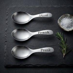 May include: Three silver-toned metal spoons with short, wide handles. Each spoon features the name Frank engraved on the handle in a different font style, ranging from a cursive script to a bold, block-style serif typeface.