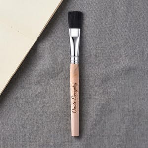 May include: A flat paint brush with black synthetic bristles, a silver metal ferrule, and a light-colored wooden handle. The handle features the text "Create Everyday" engraved in a dark, cursive script along the center of the wood.