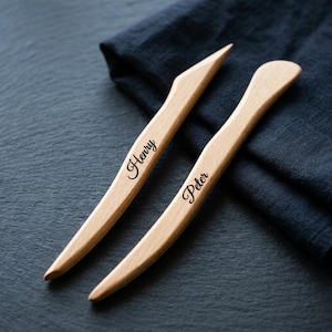 May include: Two light-colored wooden bone folders with smooth, curved edges. One tool is engraved with the name Henry in a black script font, and the other tool is engraved with the name Peter in the same black script font style.